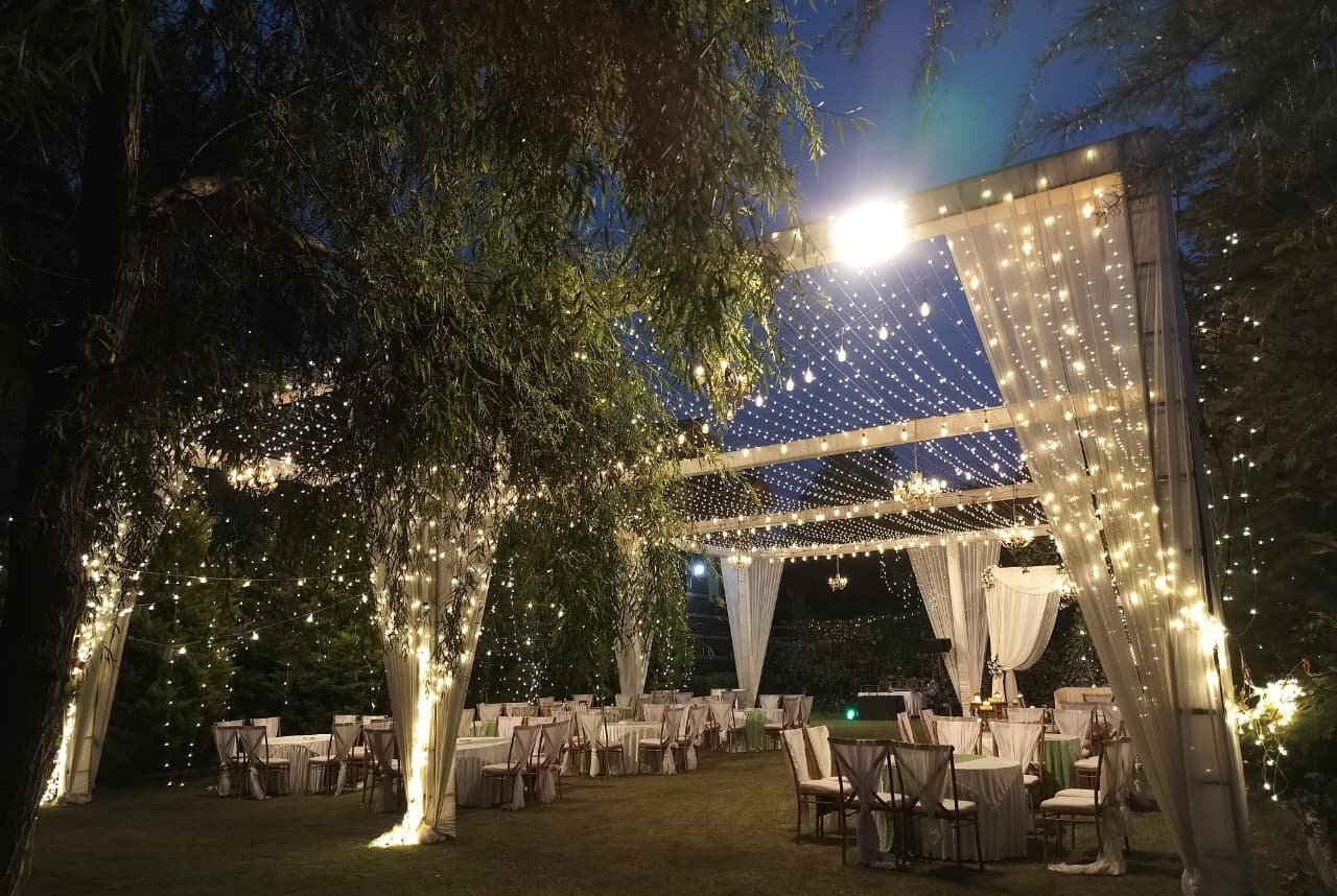 Fairy-light canopy with white drapes and chandeliers over dinner tables on the lawn at Shree Ganga Cottages, Raison
