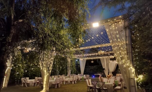 Ring ceremony setup at dusk with fairy lights, draped pillars and round tables on the lawn of Shree Ganga Cottages, Raison Kullu