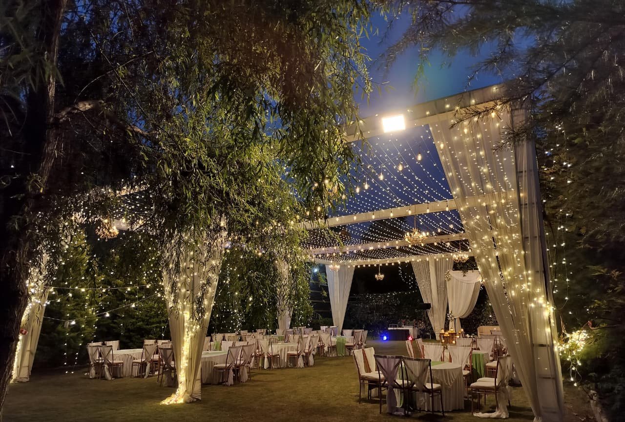 Ring ceremony setup at dusk with fairy lights, draped pillars and round tables on the lawn of Shree Ganga Cottages, Raison Kullu